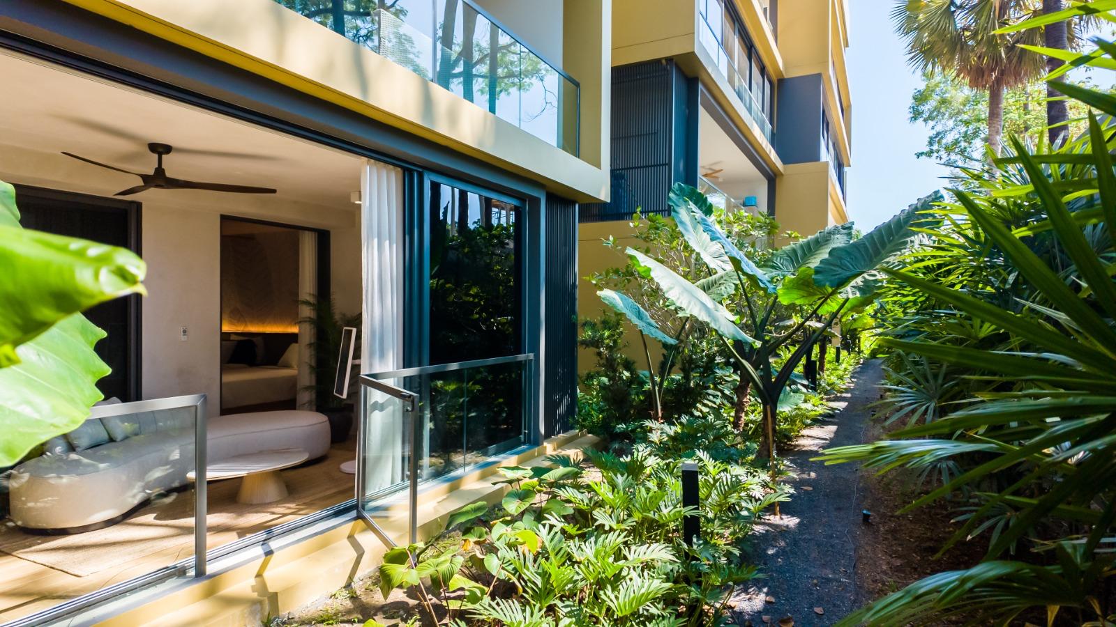 Ground-floor hospitality suite with glass walls open to a lush tropical garden path and exterior greenery outside the living area.
