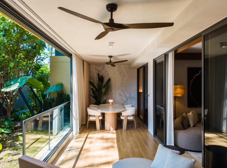 Sunlit dining area at the far end of a glass-walled corridor, a round wooden table with white chairs, and a view of lush greenery through sliding doors on the left.
