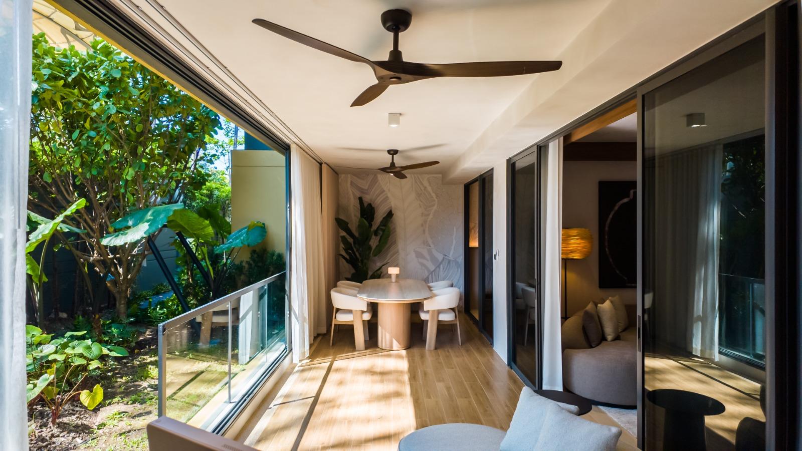 Sunlit dining area at the far end of a glass-walled corridor, a round wooden table with white chairs, and a view of lush greenery through sliding doors on the left.