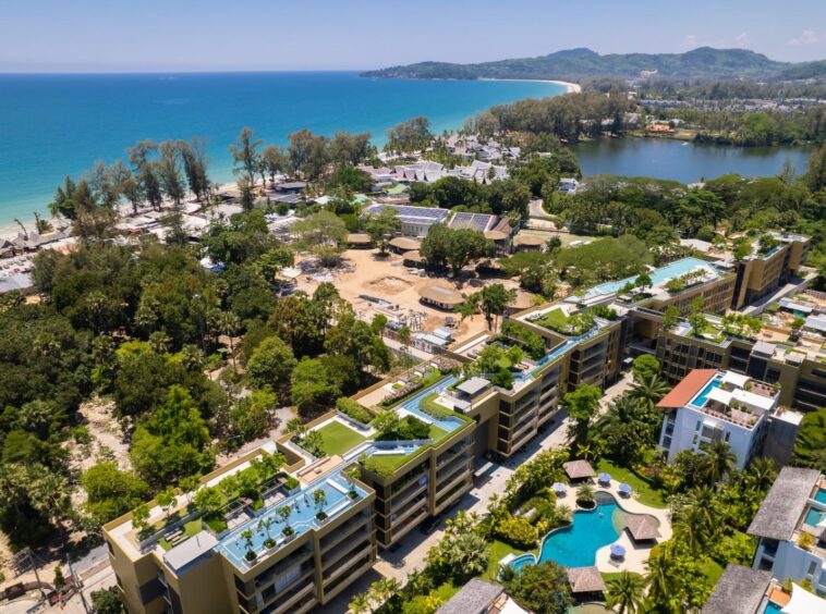 Aerial view of a coastal resort: sandy beach, turquoise sea, dense trees, and modern apartment buildings with pools nearby.
