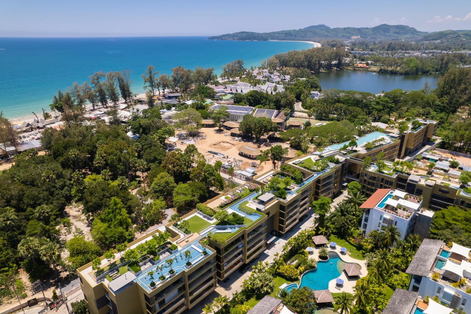Aerial view of a coastal resort: sandy beach, turquoise sea, dense trees, and modern apartment buildings with pools nearby.