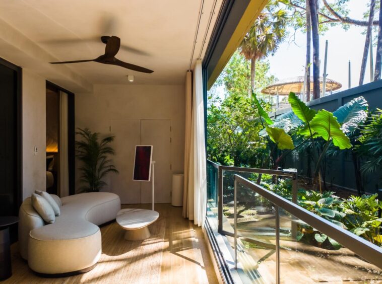 Interior living area with curved white sofa, round coffee table, and floor-to-ceiling glass doors opening to a tropical garden.