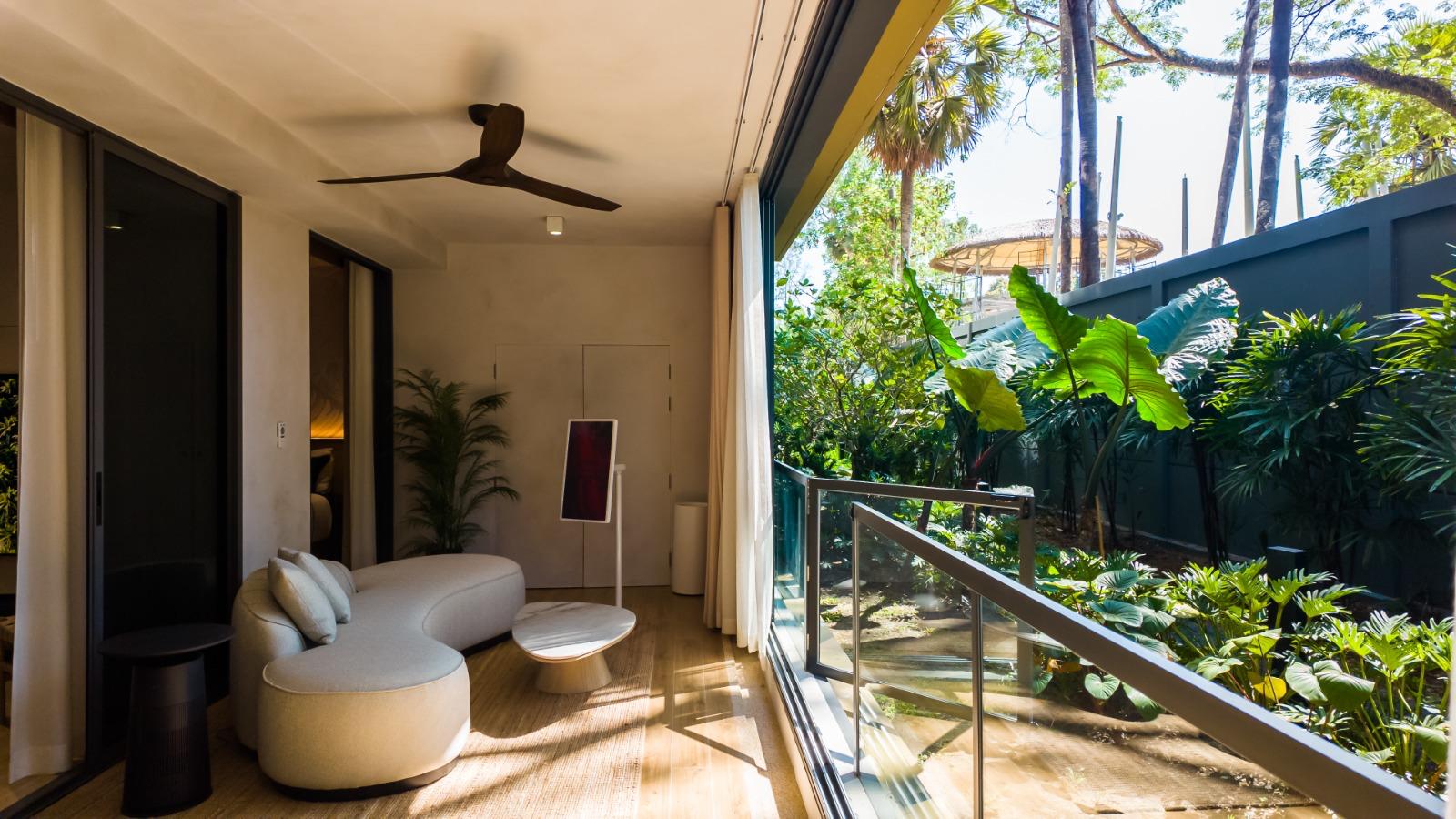 Interior living area with curved white sofa, round coffee table, and floor-to-ceiling glass doors opening to a tropical garden.