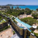 Aerial view of a coastal resort with terraced buildings, rooftop pools, and lush greenery overlooking a turquoise sea and sandy beach.
