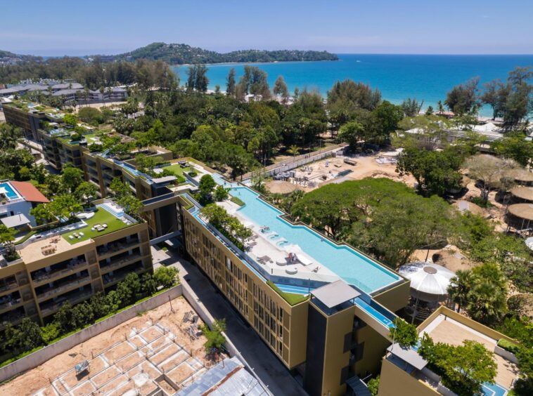 Aerial view of a coastal resort with terraced buildings, rooftop pools, and lush greenery overlooking a turquoise sea and sandy beach.