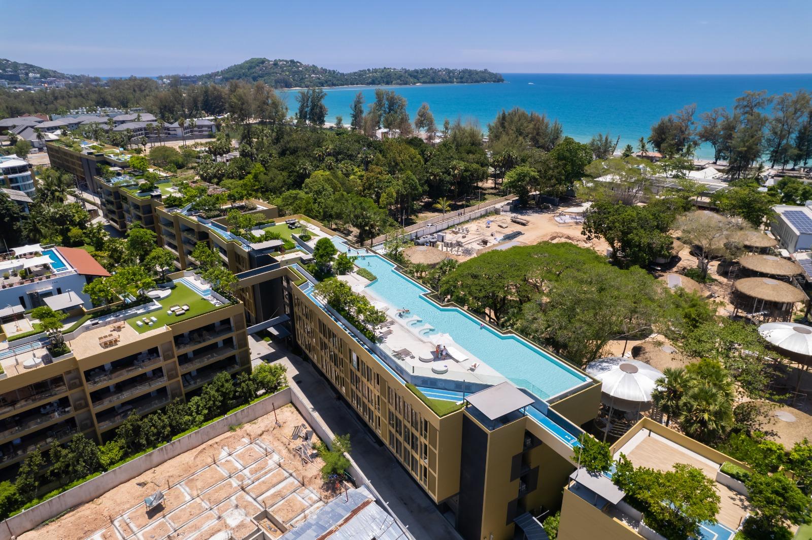 Aerial view of a coastal resort with terraced buildings, rooftop pools, and lush greenery overlooking a turquoise sea and sandy beach.