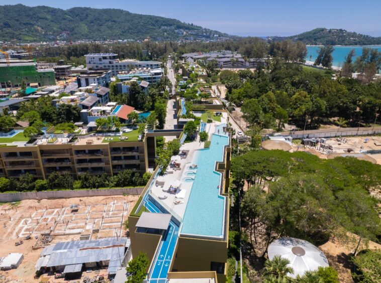 Aerial view of a modern resort with a long rooftop pool, lounge chairs, and surrounding buildings, with trees and a turquoise sea in the distance.