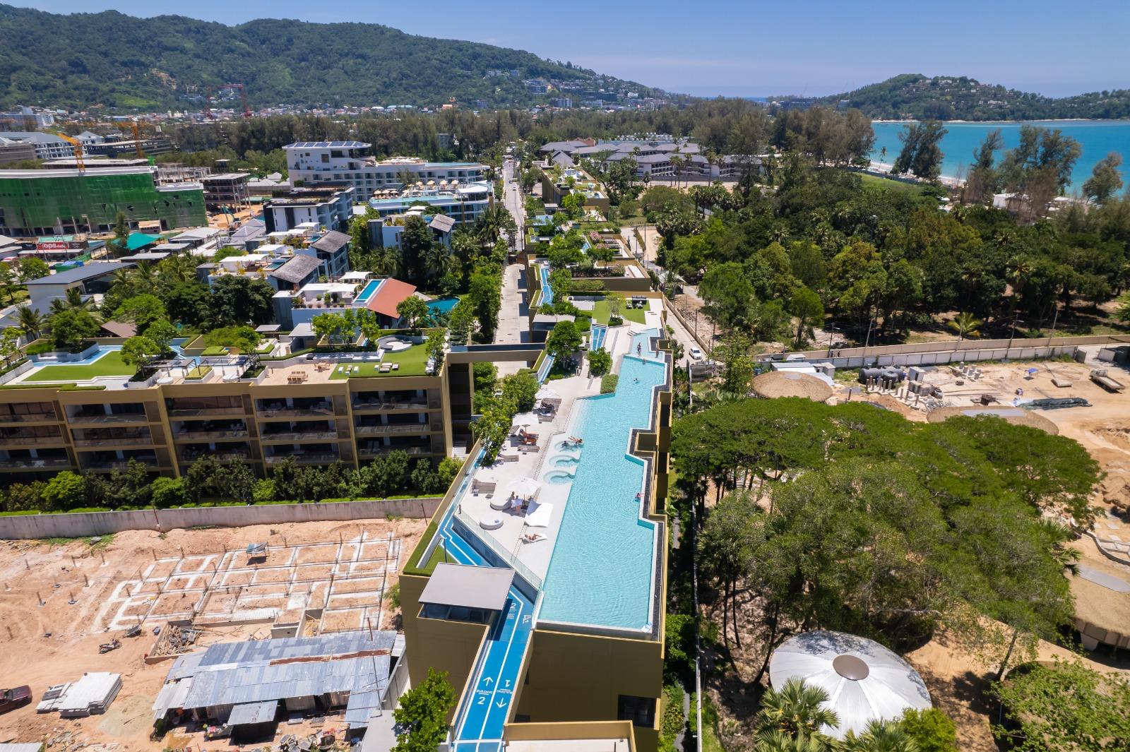Aerial view of a modern resort with a long rooftop pool, lounge chairs, and surrounding buildings, with trees and a turquoise sea in the distance.