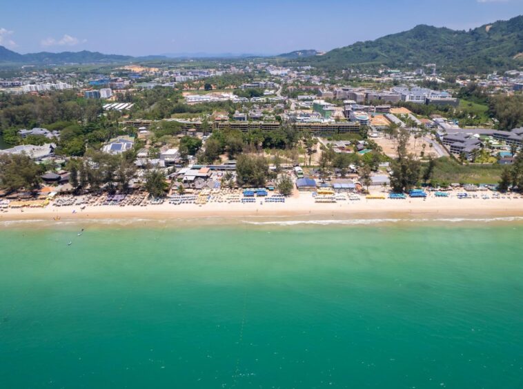 Aerial view of a tropical beach town with white sand, rows of umbrellas, and turquoise water backed by green hills and buildings.