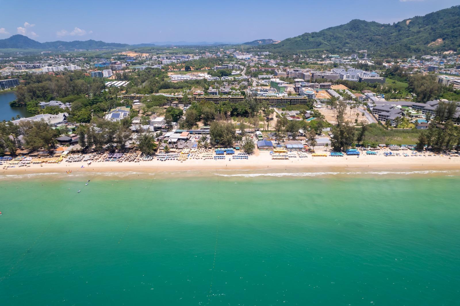 Aerial view of a tropical beach town with white sand, rows of umbrellas, and turquoise water backed by green hills and buildings.