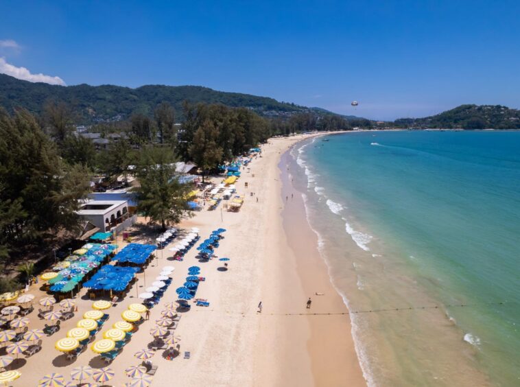 Aerial view of a sandy beach lined with yellow and blue umbrellas, turquoise sea, and tree-covered hills in the background.