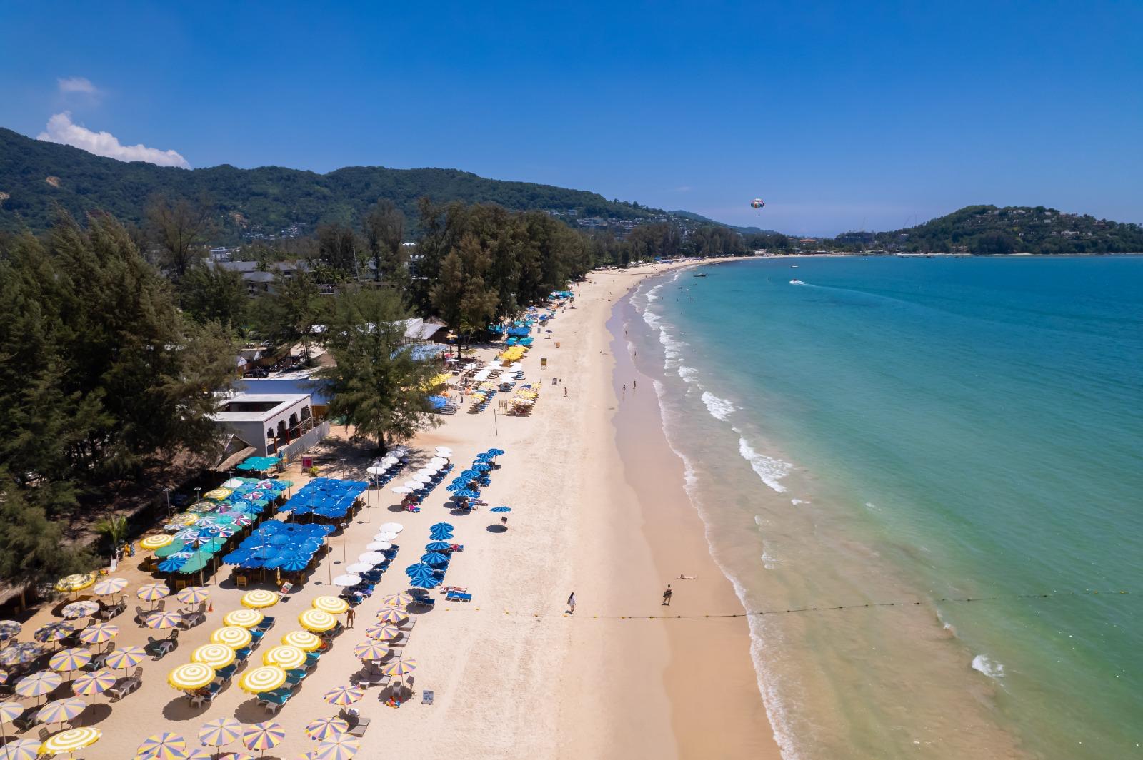 Aerial view of a sandy beach lined with yellow and blue umbrellas, turquoise sea, and tree-covered hills in the background.