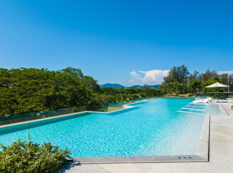 Long rectangular pool with bright blue water, lounge chairs and white umbrellas along a sunlit deck, green trees in the background.