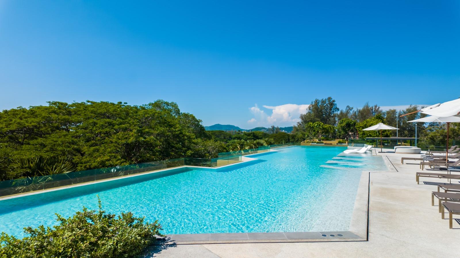 Long rectangular pool with bright blue water, lounge chairs and white umbrellas along a sunlit deck, green trees in the background.
