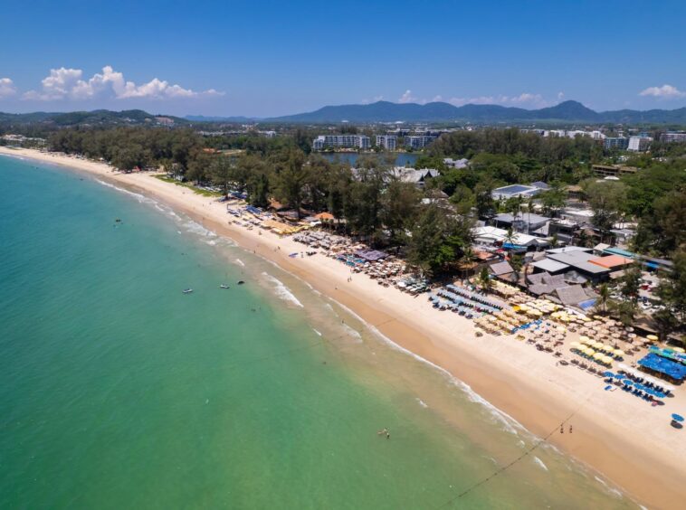 Aerial view of a tropical beach with turquoise water, a long sandy shore, and rows of colorful umbrellas along the coast backed by trees and buildings.