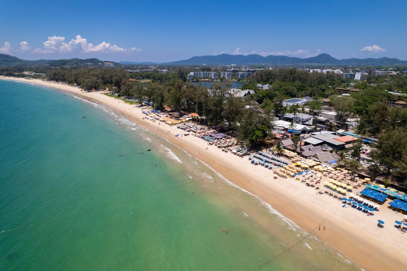 Aerial view of a tropical beach with turquoise water, a long sandy shore, and rows of colorful umbrellas along the coast backed by trees and buildings.