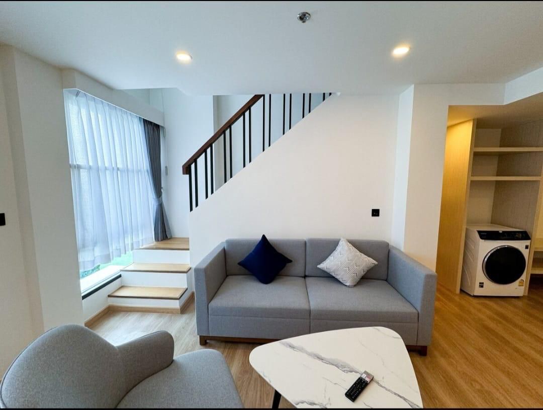 Bright modern living area with a gray sofa, two throw pillows, a white marble coffee table, and a staircase with black railing in the background; large window with sheer curtains along the left wall.