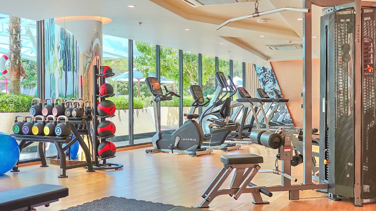 Gym interior with cardio machines (treadmills, bikes) facing large windows, plus kettlebells, medicine balls on racks and weight stack cable machine on the right.