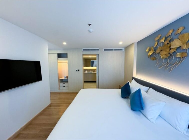 Modern hotel bedroom with a large bed, blue pillows, wall-mounted TV, and gold leaf wall sculpture above the headboard.