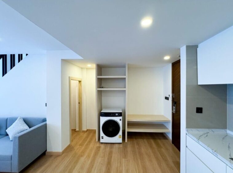 Open-concept living area with a front-loading washing machine under built-in shelves and a wooden bench to the right, in a bright space.