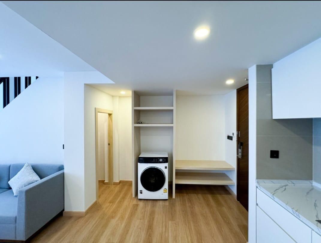 Open-concept living area with a front-loading washing machine under built-in shelves and a wooden bench to the right, in a bright space.