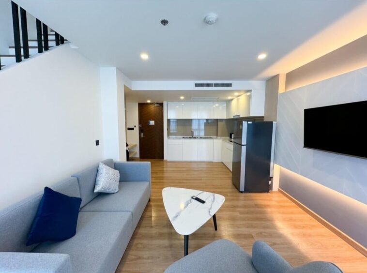 Open-plan living area with a gray sofa, marble coffee table, and a wall-mounted TV; kitchen with white cabinets and a stainless fridge in the background.