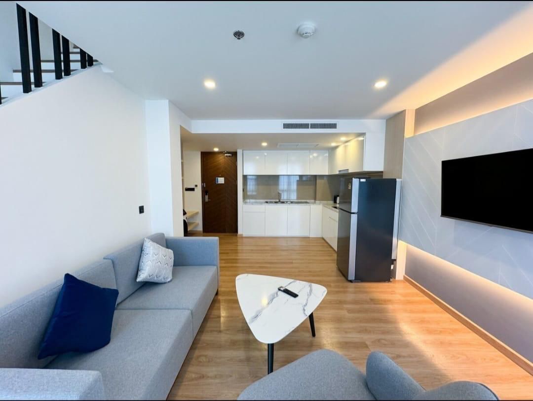 Open-plan living area with a gray sofa, marble coffee table, and a wall-mounted TV; kitchen with white cabinets and a stainless fridge in the background.