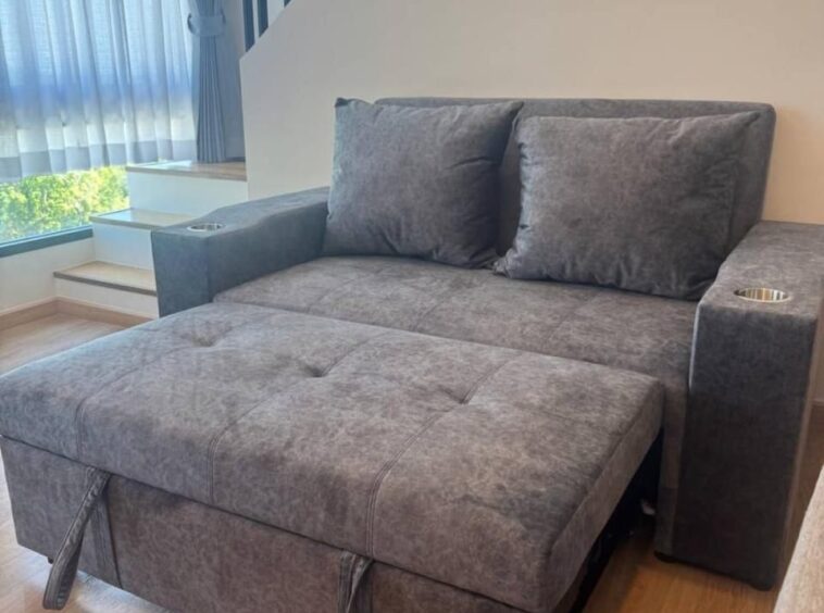 Gray fabric sofa with a chaise lounge and two back cushions in a bright living room by a large window with blue curtains.