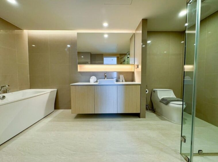 A contemporary bathroom with a freestanding white bathtub on the left, a wood vanity with sink and mirror in the center, and a toilet on the right behind a glass shower wall.