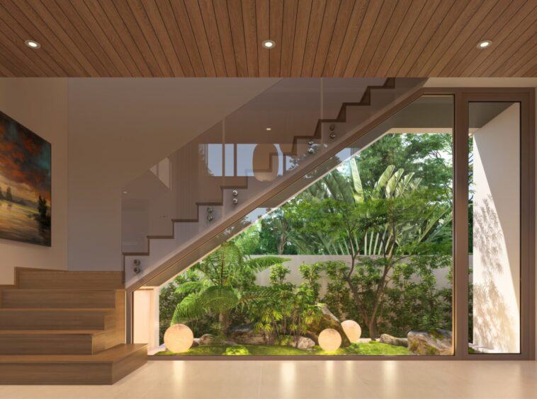 Modern interior with wooden staircase leading to a glass-walled courtyard garden visible through large windows and a glass railing reflecting greenery above- staircase wall art on left; warm wood ceiling panels.