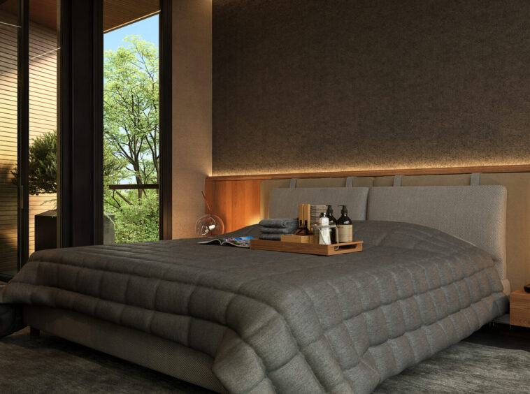 Modern bedroom with a low, quilted gray bed, wooden headboard, and glass doors showing greenery outside.