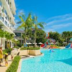 Family walks along a bright blue resort pool edge with palm trees and colorful water slides in the background under a sunny sky.
