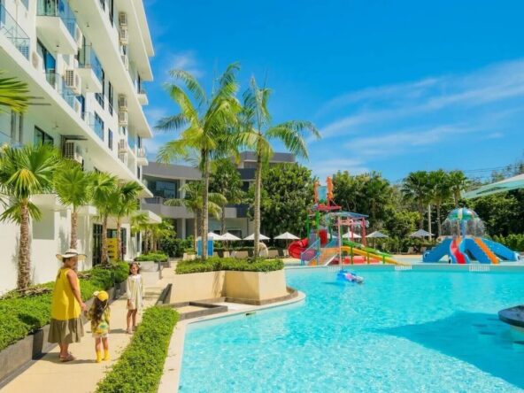 Family walks along a bright blue resort pool edge with palm trees and colorful water slides in the background under a sunny sky.