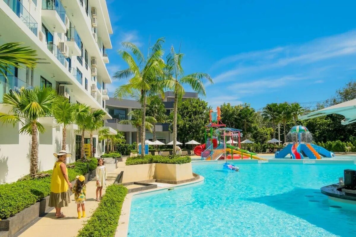 Family walks along a bright blue resort pool edge with palm trees and colorful water slides in the background under a sunny sky.