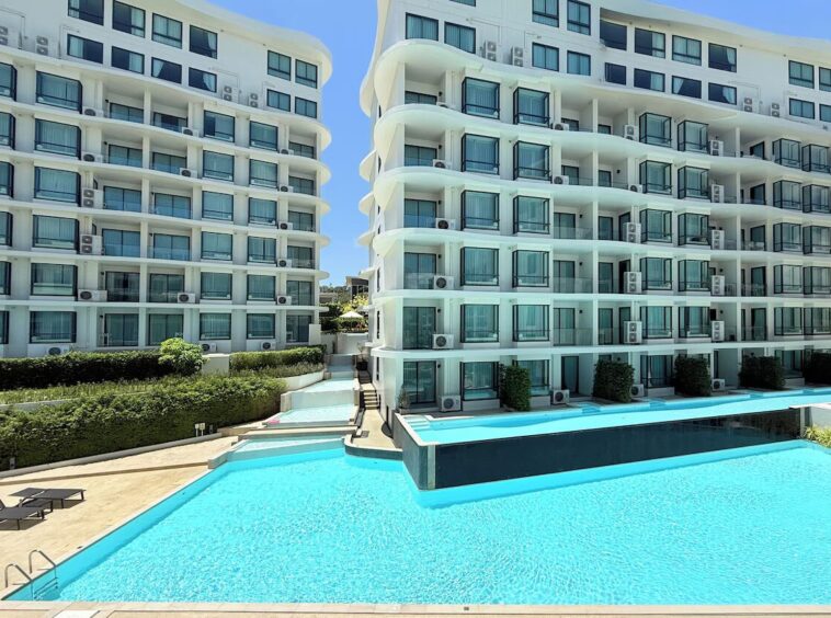 Two white modern apartment buildings flank a bright blue swimming pool with lounge chairs nearby.