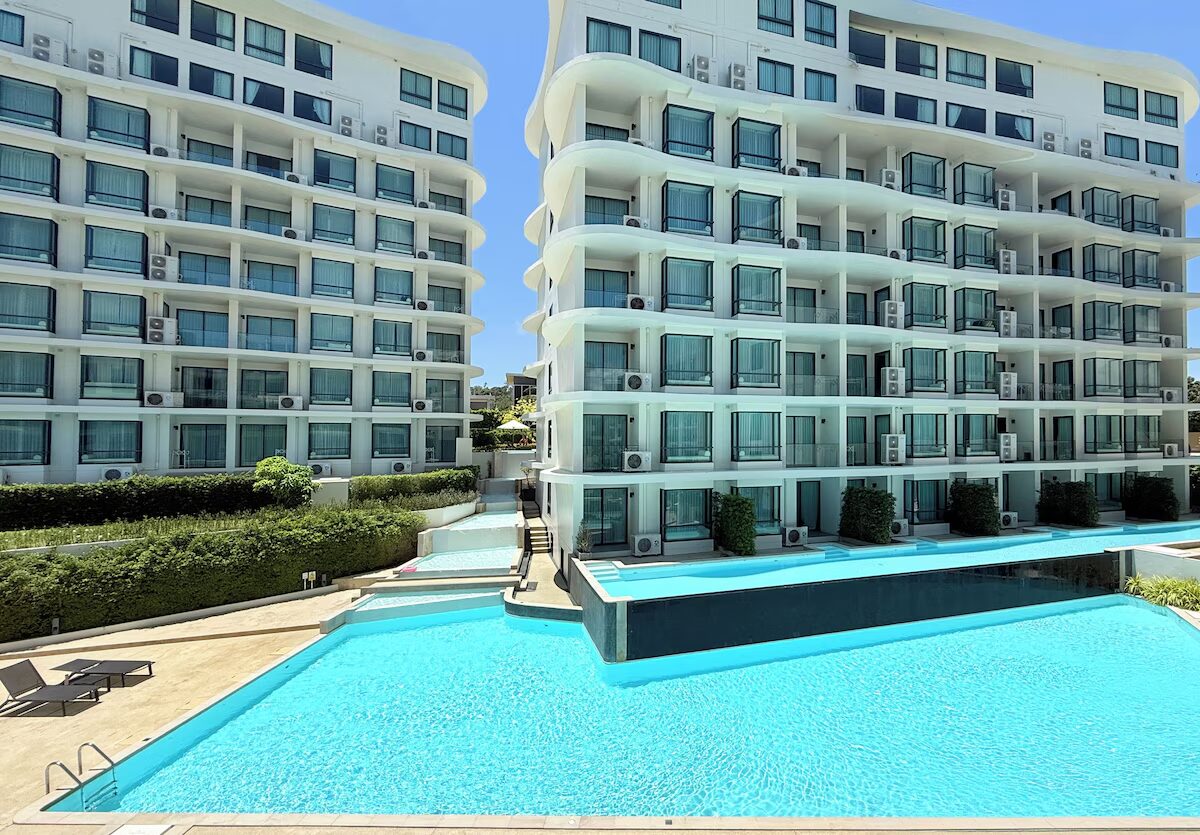 Two white modern apartment buildings flank a bright blue swimming pool with lounge chairs nearby.