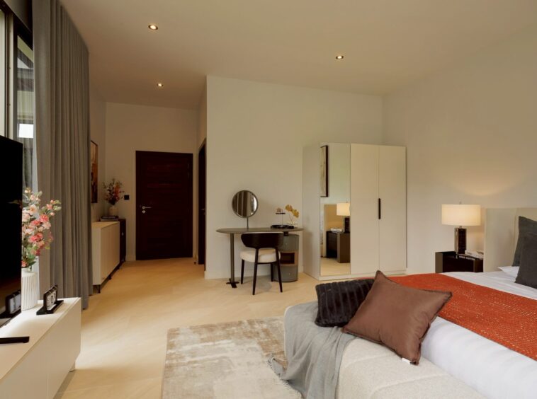 Modern hotel bedroom with a large bed, orange throw, brown pillows, and a bedside lamp on the right.