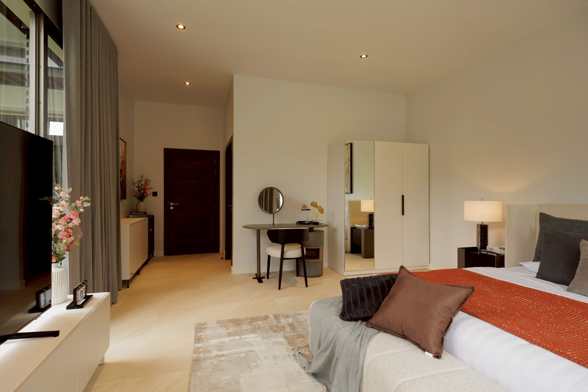 Modern hotel bedroom with a large bed, orange throw, brown pillows, and a bedside lamp on the right.