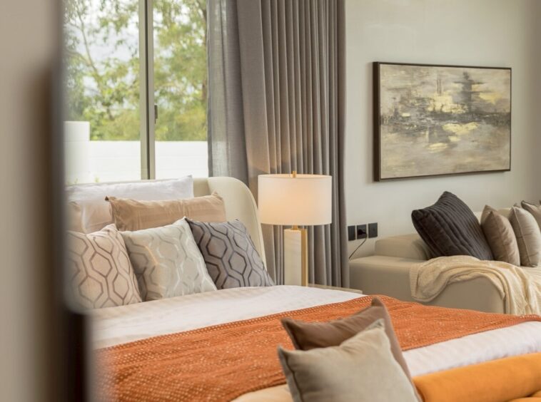 Sunlit hotel-style bedroom with white bedding, orange throw, and gray patterned pillows at the foot of the bed.