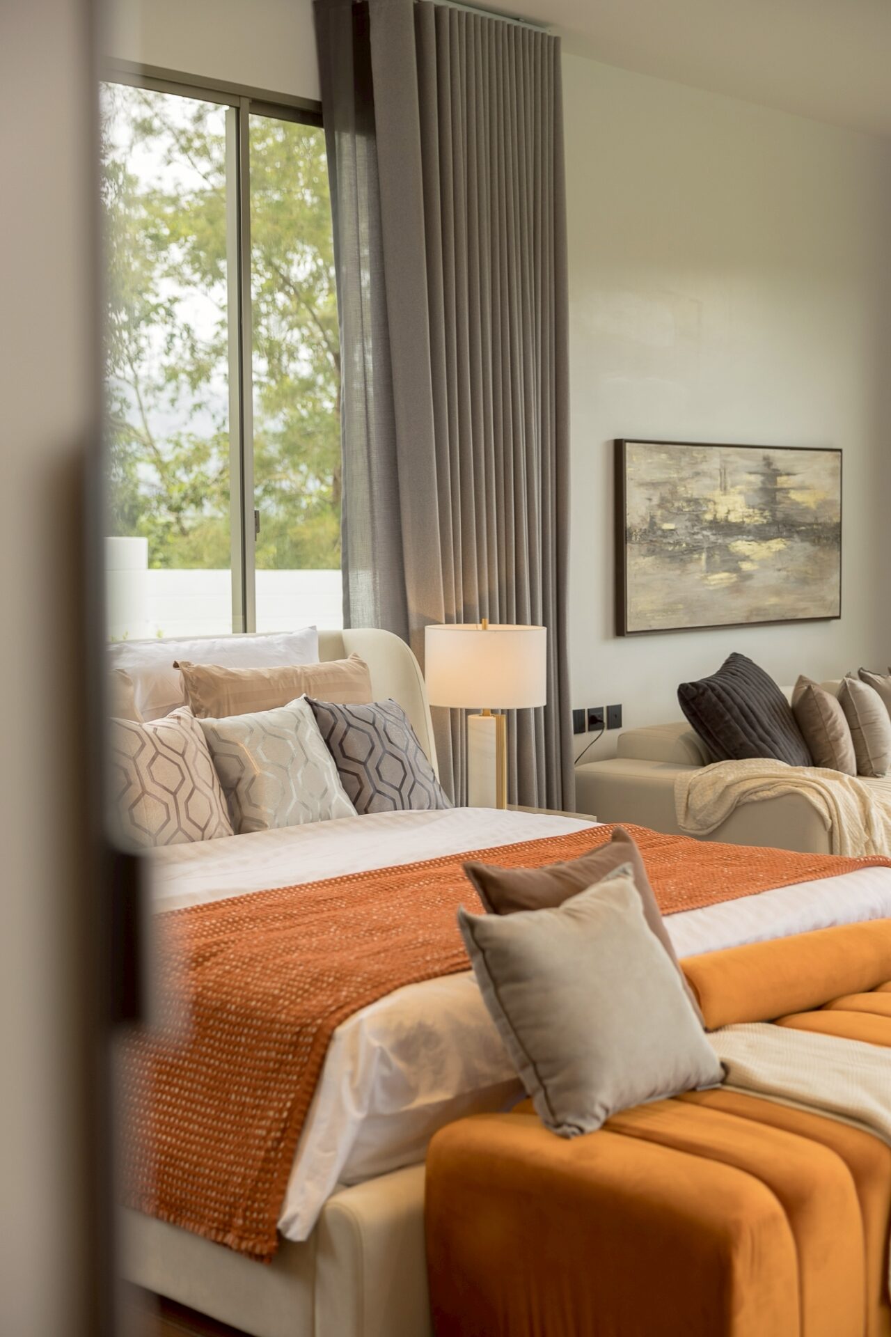 Sunlit hotel-style bedroom with white bedding, orange throw, and gray patterned pillows at the foot of the bed.
