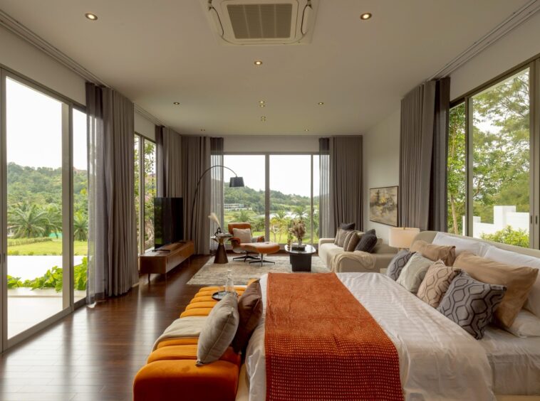 Spacious modern bedroom/living area with floor-to-ceiling glass walls, neutral furniture, and a view of trees and greenery outside.