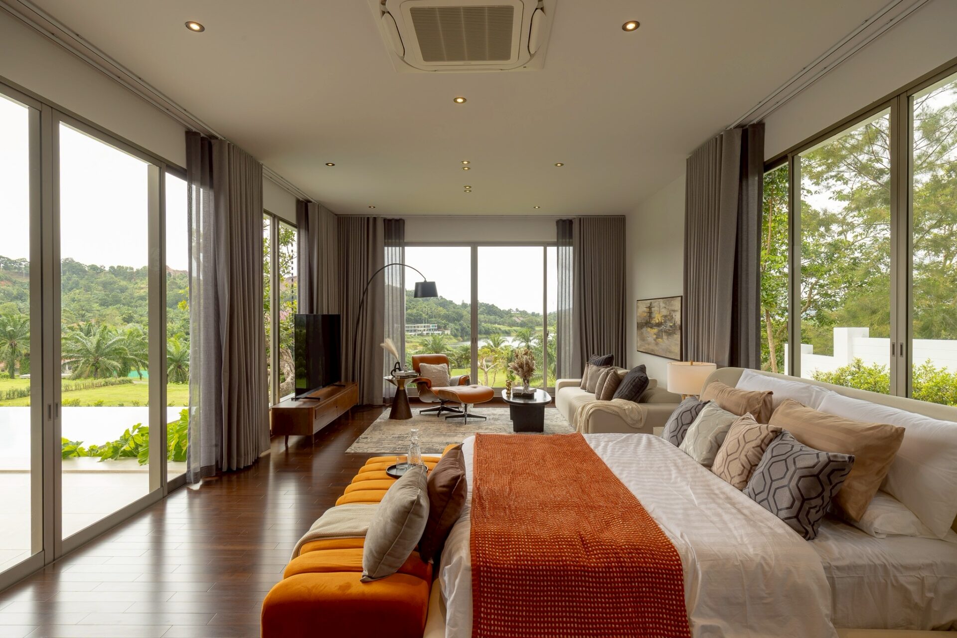 Spacious modern bedroom/living area with floor-to-ceiling glass walls, neutral furniture, and a view of trees and greenery outside.
