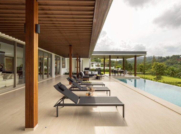 Modern poolside patio with black lounge chairs under a wooden overhang and glass-walled house exterior.