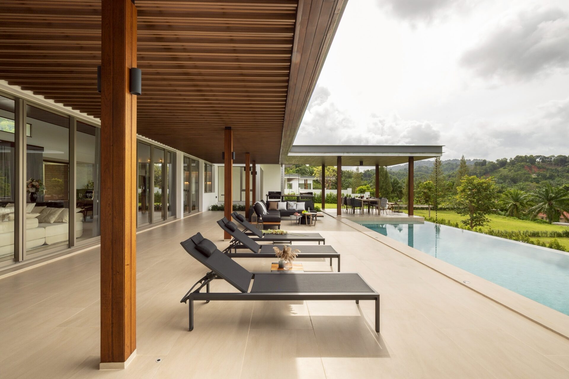 Modern poolside patio with black lounge chairs under a wooden overhang and glass-walled house exterior.