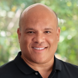 Head-and-shoulders portrait of a smiling man with a bald head wearing a black polo shirt, outdoors with a blurred green background