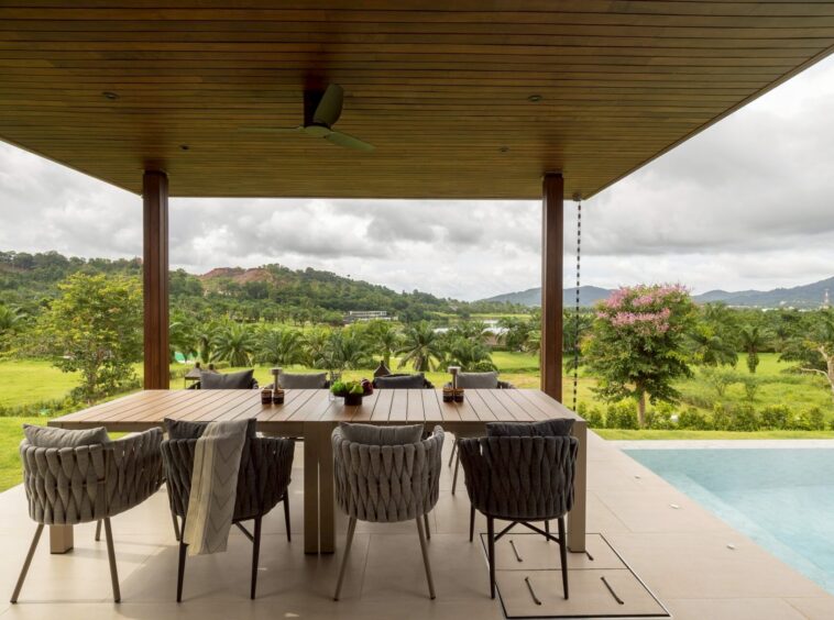 Outdoor covered dining area with a long wooden table and woven chairs, overlooking a lush landscape of trees and hills by a poolside patio.