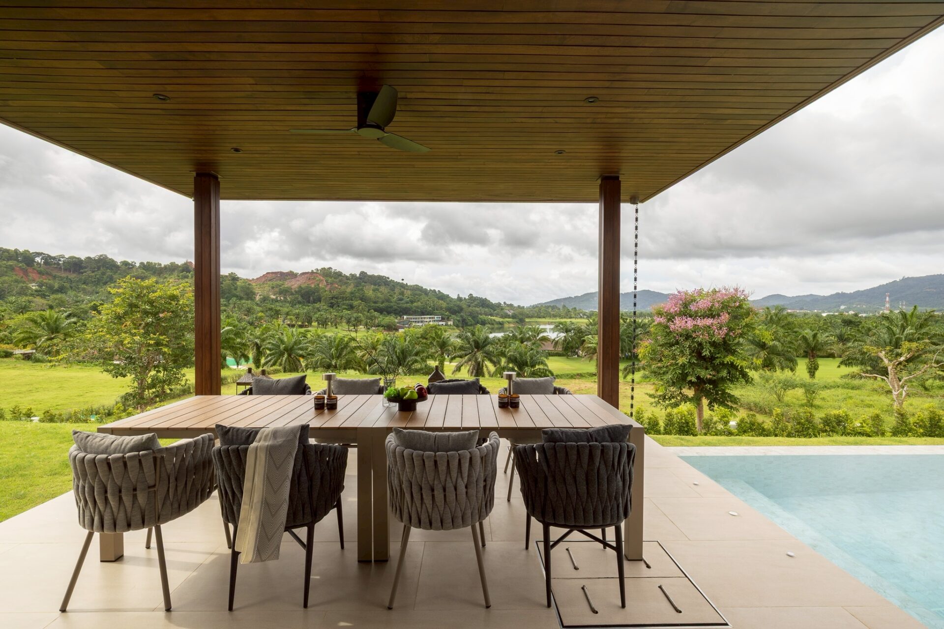 Outdoor covered dining area with a long wooden table and woven chairs, overlooking a lush landscape of trees and hills by a poolside patio.