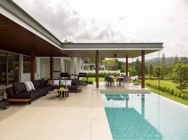 Modern outdoor living area with beige tiled patio, black sectional sofa, and a rectangular pool full of crystal water.