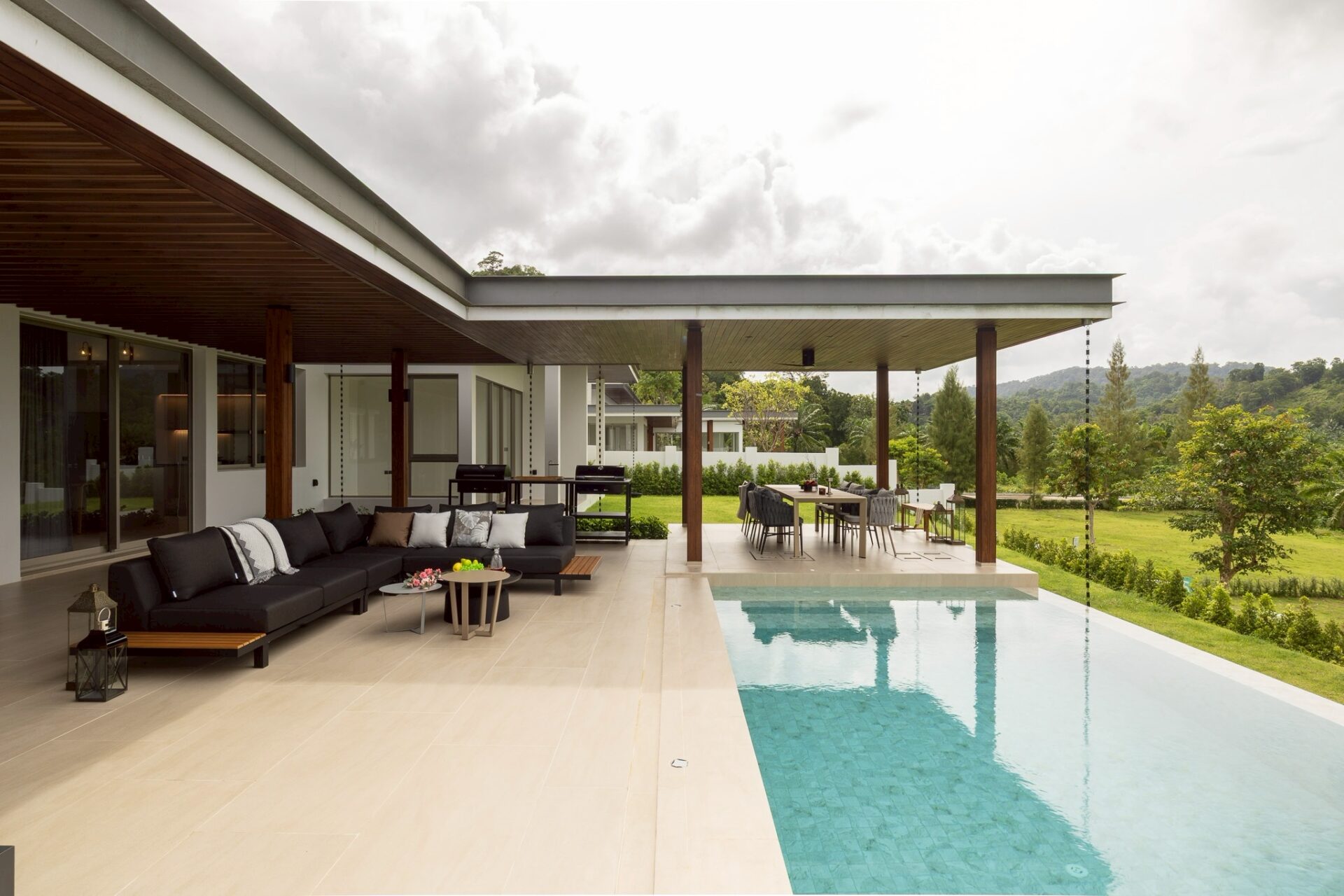 Modern outdoor living area with beige tiled patio, black sectional sofa, and a rectangular pool full of crystal water.