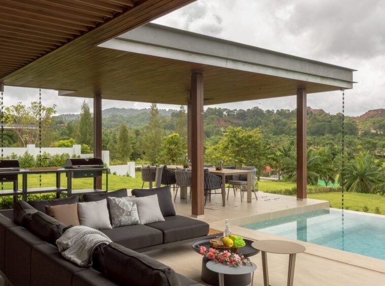 Outdoor covered patio with a dark sectional sofa, coffee tables, and a rectangular pool, overlooking lush green hills.